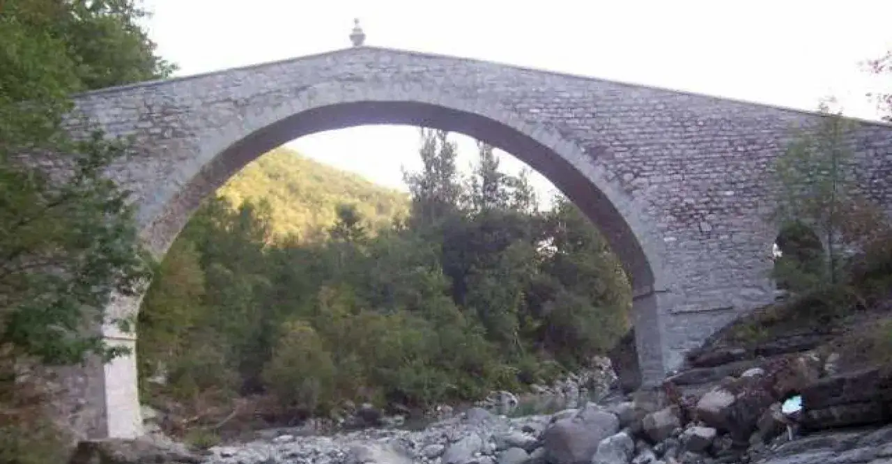 Antico ponte in pietra, simile a un ponte tibetano, attraversa un torrente in un paesaggio verdeggiante dell'Emilia Romagna.