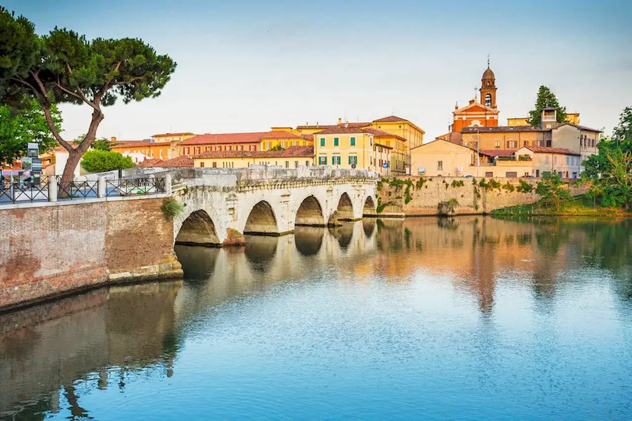 Il Ponte di Tiberio a Rimini, con i suoi archi maestosi, si specchia nelle acque tranquille del fiume, circondato da edifici storici e un cielo limpido.