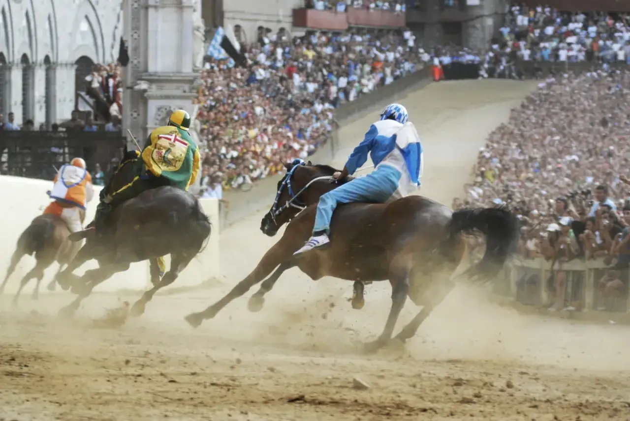 Cavalli e fantini in gara durante il Palio dell'Assunta a Siena, tra la polvere sollevata e la folla festante.