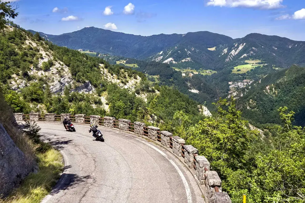 Due motociclisti affrontano una curva su una strada panoramica, esplorando i fantastici itinerari moto Emilia Romagna tra montagne verdi e cielo azzurro.