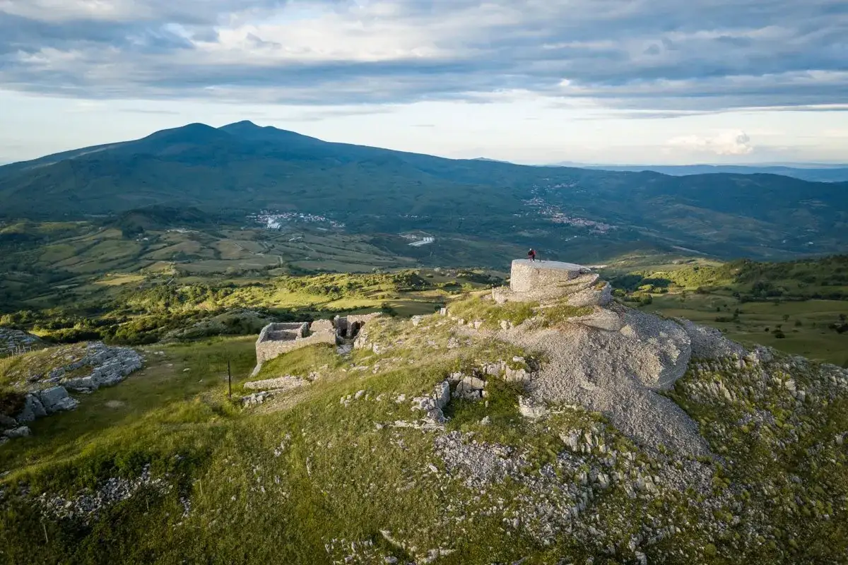 Rovine antiche su un crinale erboso, con una persona che ammira il panorama del monte Labbro in lontananza.