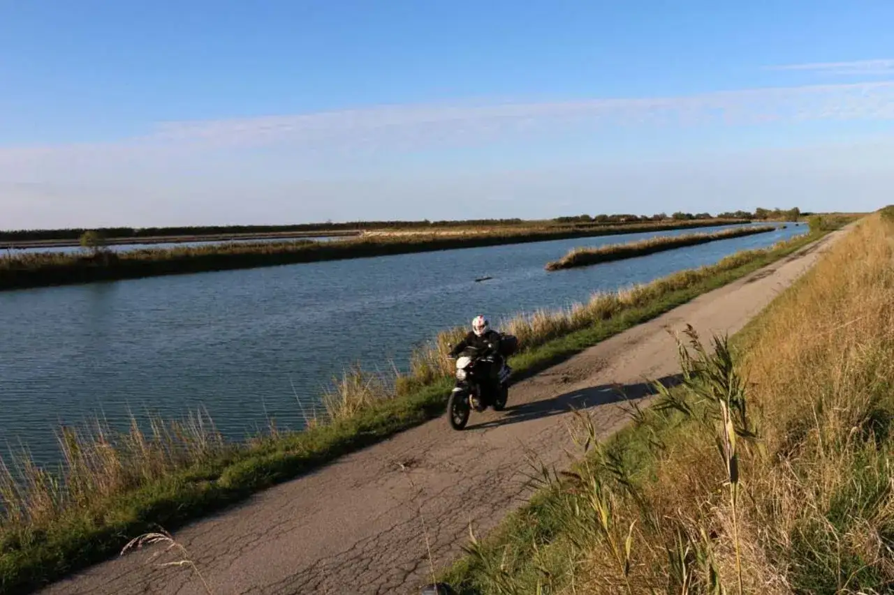 Un motociclista esplora il Delta del Po, percorrendo una strada sterrata tra canali e vegetazione rigogliosa sotto un cielo azzurro.