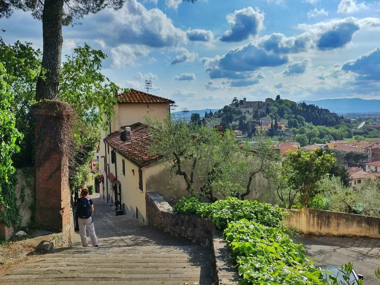 Una persona con zaino scende una scalinata in pietra, godendosi il panorama di Monte Morello e delle colline toscane.