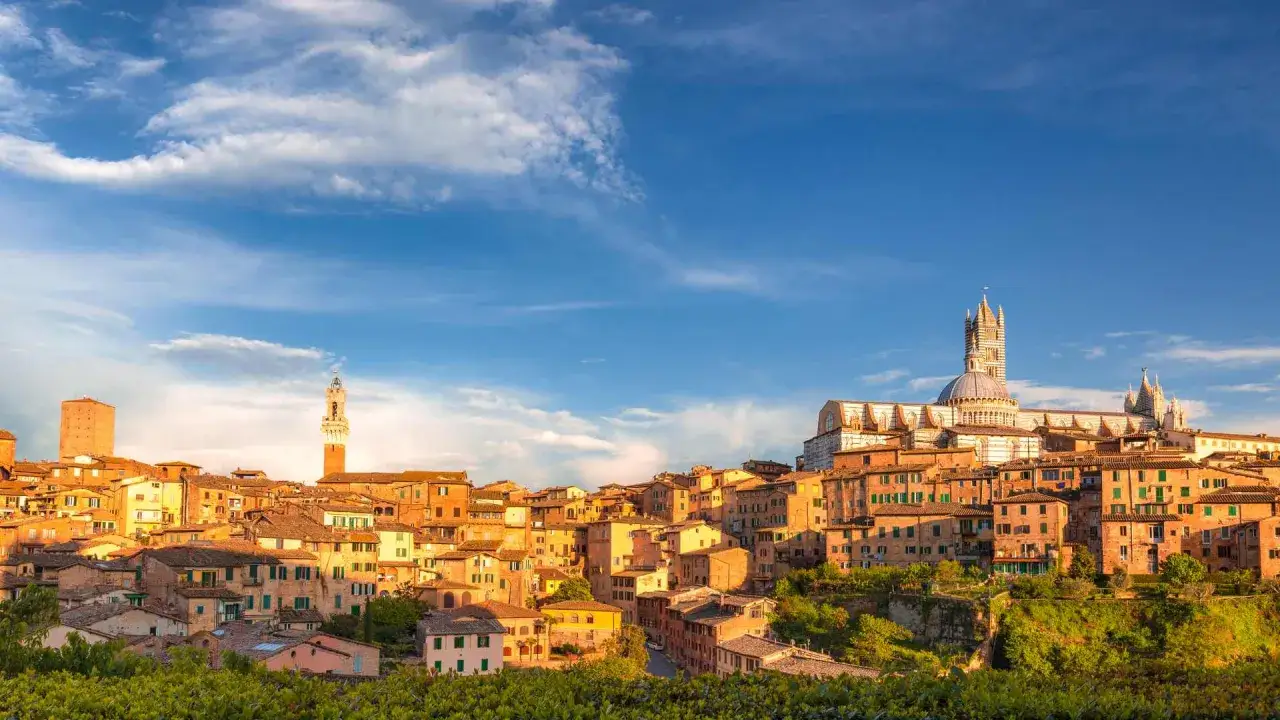 Siena da vedere: panorama di tetti in cotto, torri medievali e il Duomo sotto un cielo azzurro con nuvole bianche.