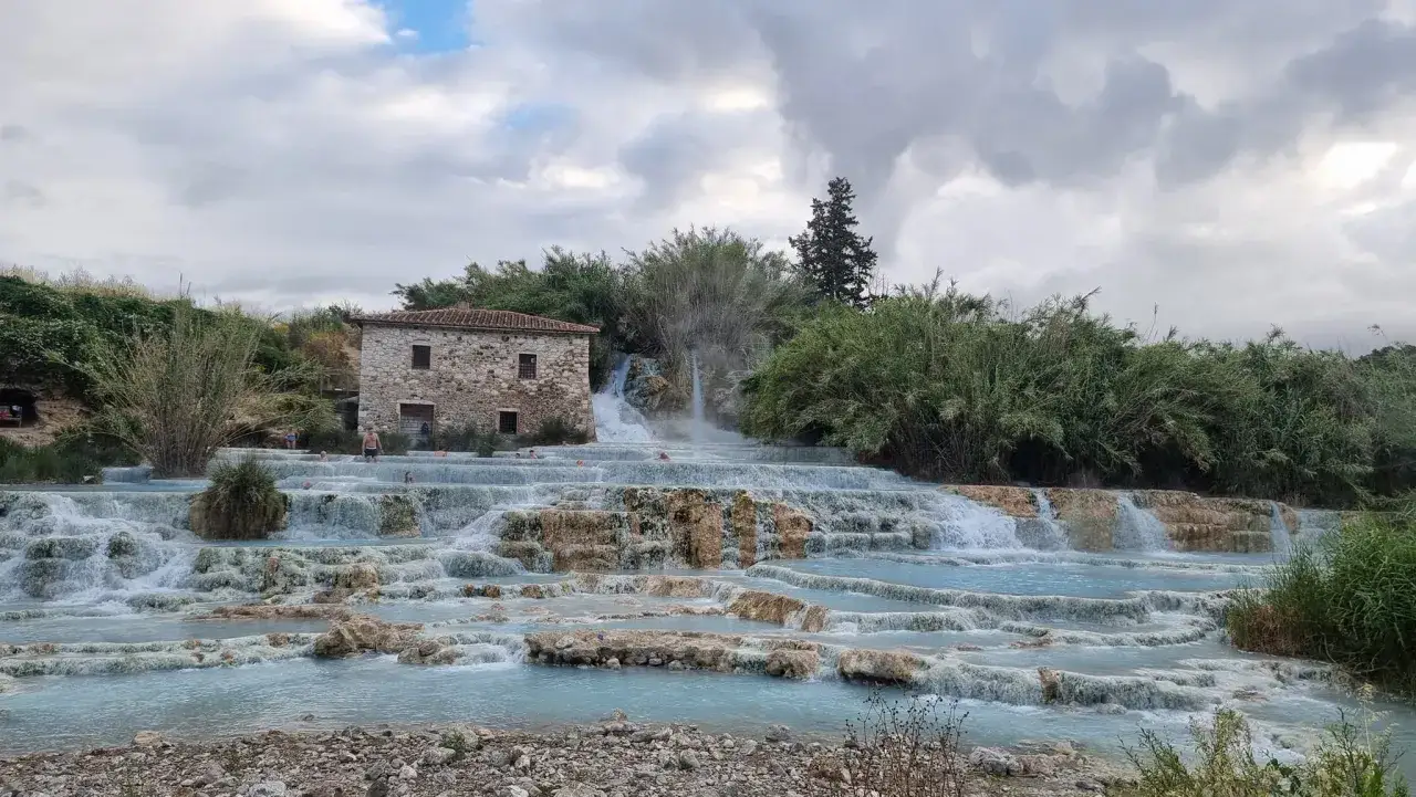 Cascate naturali in Toscana con un antico casale in pietra. L'acqua turchese scorre su rocce calcaree, creando piscine termali dove alcune persone si rilassano.