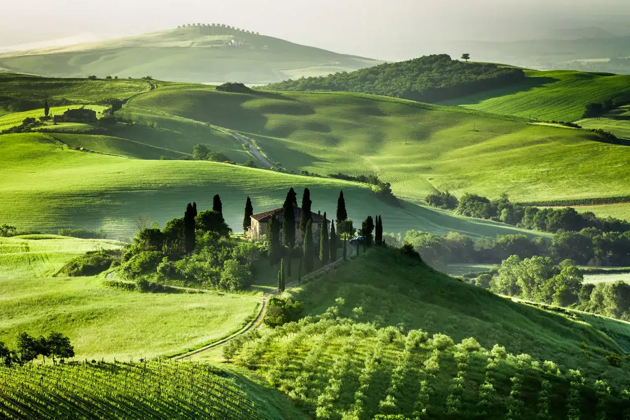 Colline verdi ondulate in Toscana, con una casa colonica e cipressi in primo piano, evocano la pace dei borghi vicino Montalcino.