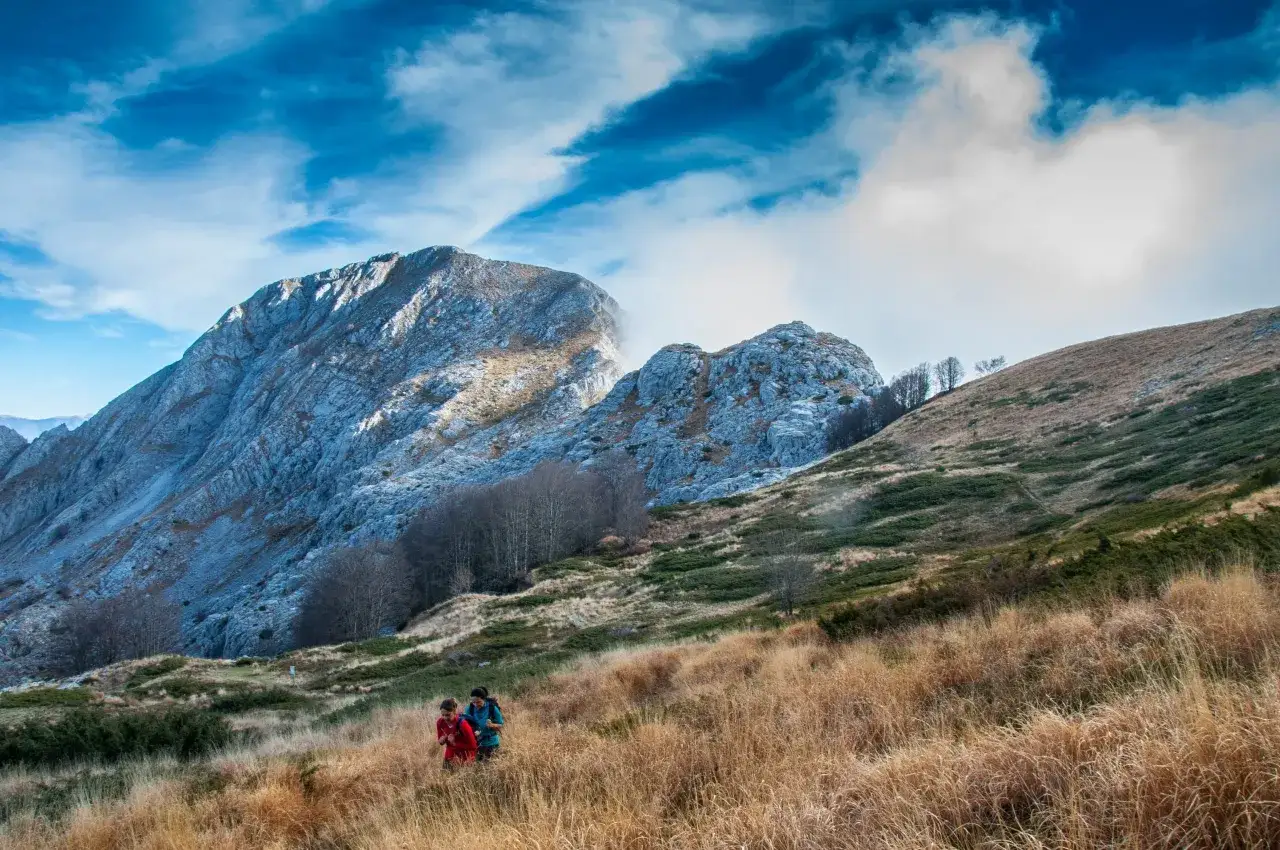 Due escursionisti attraversano un prato dorato, con le maestose Alpi Apuane sullo sfondo, un'esperienza di trekking tra i più belli.