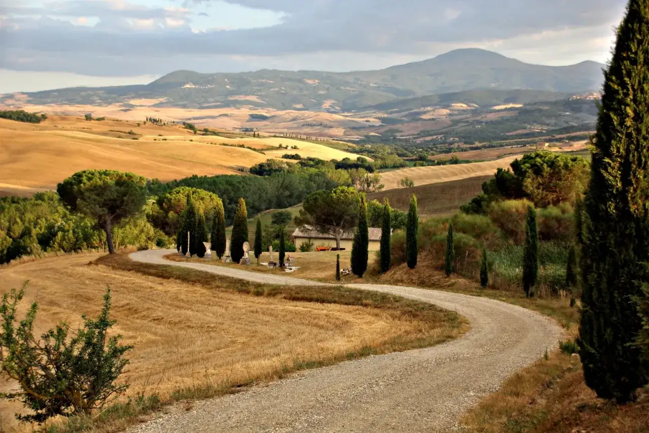 Strada sterrata serpeggia tra colline dorate e cipressi, un invito a percorrere la via Francigena Toscana verso un casolare solitario.