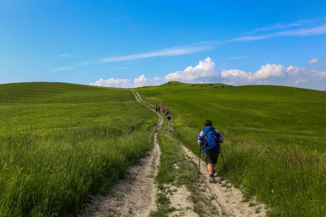 Un gruppo di escursionisti percorre un sentiero sterrato attraverso dolci colline verdi, sotto un cielo azzurro con nuvole bianche. Un'avventura lungo la via Lauretana Toscana.