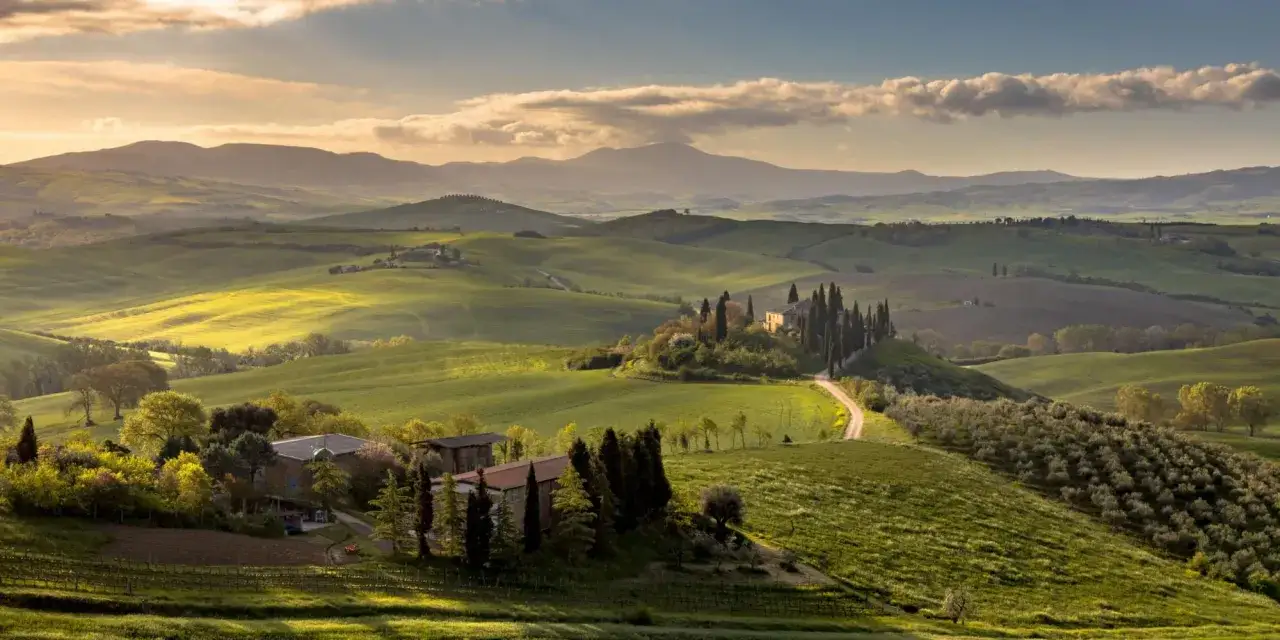Dolci colline senesi illuminate dal sole del tramonto, con casali e cipressi che disegnano il paesaggio.