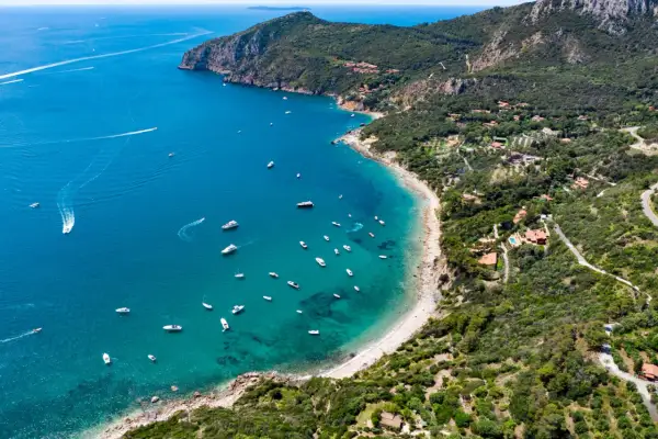 Baia con acque cristalline e numerose barche al largo della costa del Monte Argentario. Ville immerse nel verde e una strada panoramica.