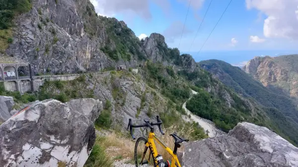 Bicicletta gialla in primo piano, con un panorama mozzafiato di montagne scoscese e un passo del vestito che si snoda tra le rocce.