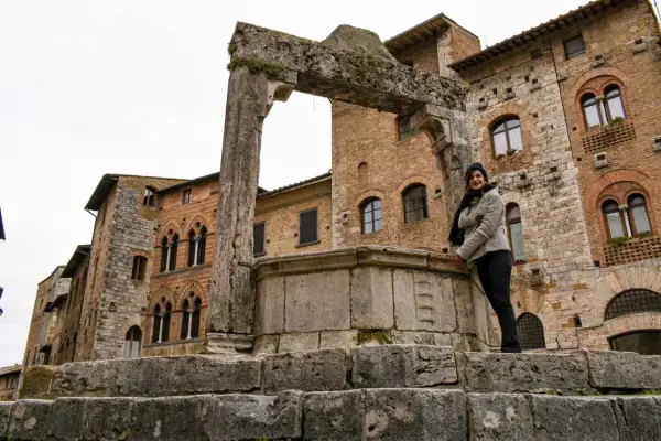 Donna sorride vicino a un antico pozzo a San Gimignano, con palazzi medievali sullo sfondo.
