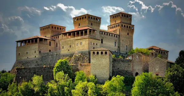 Rocche imponenti e torri medievali, tra i borghi vicino Parma, si stagliano contro un cielo nuvoloso, circondate da un verde lussureggiante.