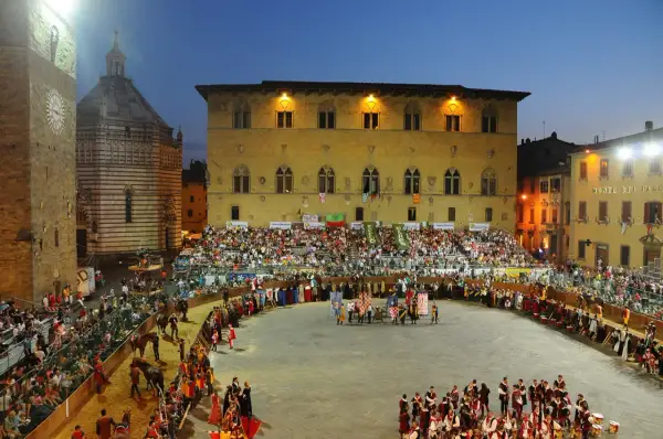 La giostra dell'orso in piazza, con figuranti in costume, cavalli e tribune gremite di spettatori sotto il cielo serale.