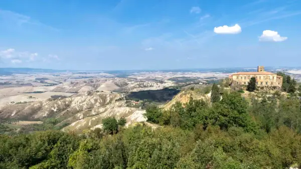 Panoramica delle balze di Volterra, con un antico palazzo in pietra su una collina e dolci colline ondulate all'orizzonte sotto un cielo azzurro.