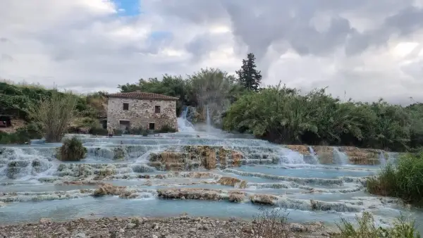 Cascate naturali in Toscana con un antico casale in pietra. L'acqua azzurra scorre su rocce calcaree, creando piscine termali dove alcune persone si rilassano.