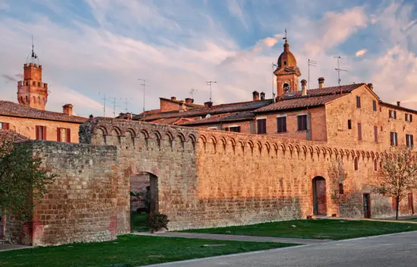 Antica cinta muraria e torri in mattoni rossi sotto un cielo azzurro striato di nuvole rosa, tipico delle colline senesi al tramonto.