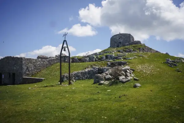 Rovine di pietra e una campana sul **monte Labbro**, sotto un cielo azzurro con nuvole bianche.