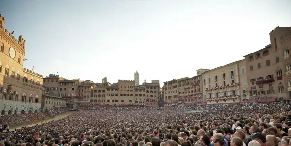 Folla immensa in Piazza del Campo a Siena, in attesa di sapere quand'è il Palio. Edifici storici e cielo terso.