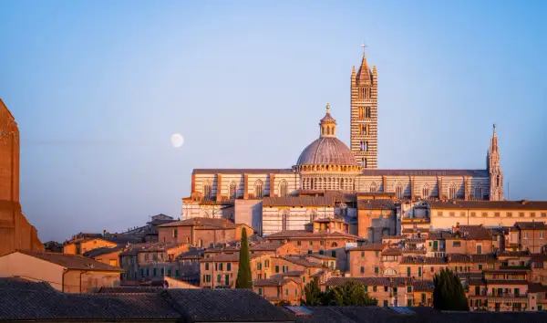 Il Duomo di Siena, con la sua torre campanaria e la cupola, domina il paesaggio urbano al tramonto. La luna piena illumina il cielo azzurro. Un'immagine che evoca la bellezza del **museum** di Siena.
