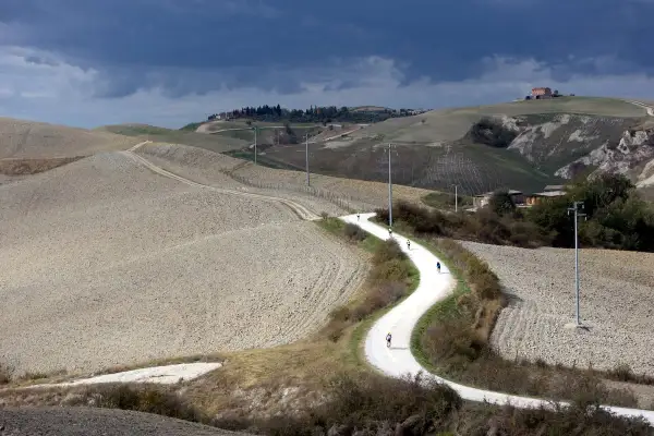 Ciclisti affrontano una strada eroica toscana, serpeggiando tra colline arate sotto un cielo minaccioso.