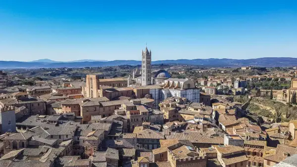 Panoramica di **Siena da vedere**: tetti in cotto, il Duomo e il Campanile svettano su un paesaggio collinare sotto un cielo azzurro.