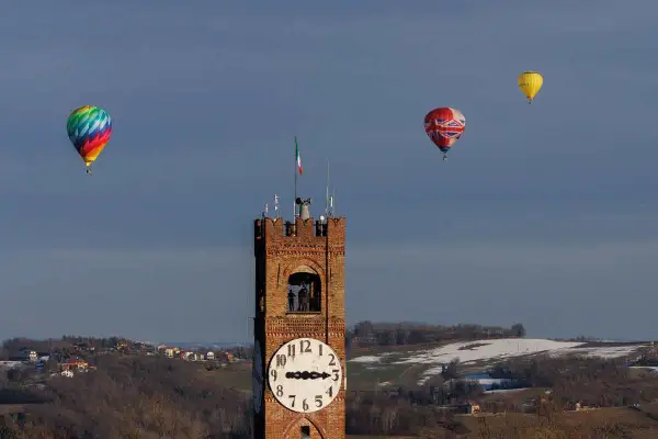 Un colorato evento mongolfiere vola nel cielo azzurro sopra una torre dell'orologio e un paesaggio collinare innevato.